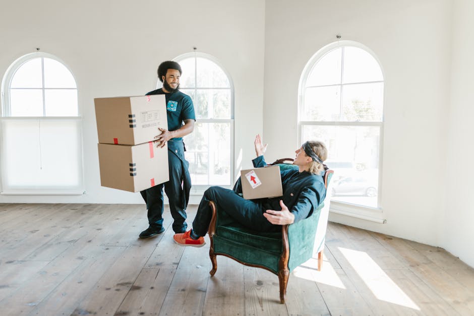 A person wearing a dark shirt and blue trousers is seen holding a large cardboard moving box marked with a red and white caution label indicating 'THIS SIDE UP.' The individual is gripping the box wit